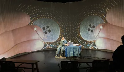 Man sitting at table on stage with projection of hands on screen behind him